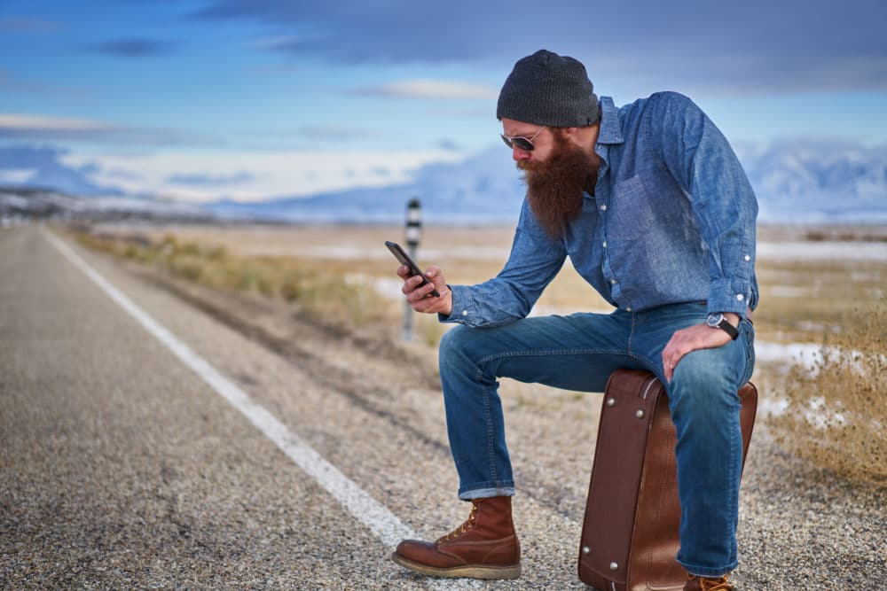 bearded hitch hiker sitting on suit case using smart phone